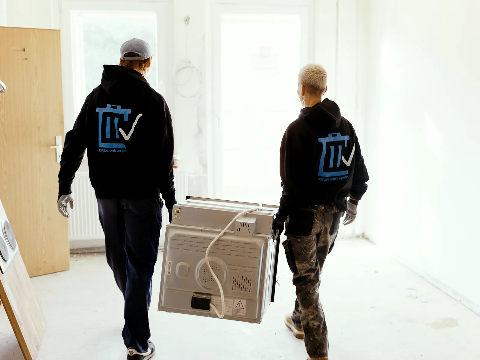 Zwei Mitarbeiter von Sorglos Entrümpeln in Hoodies mit Firmenlogo tragen einen ausgebauten Backofen durch eine leere, helle Wohnung