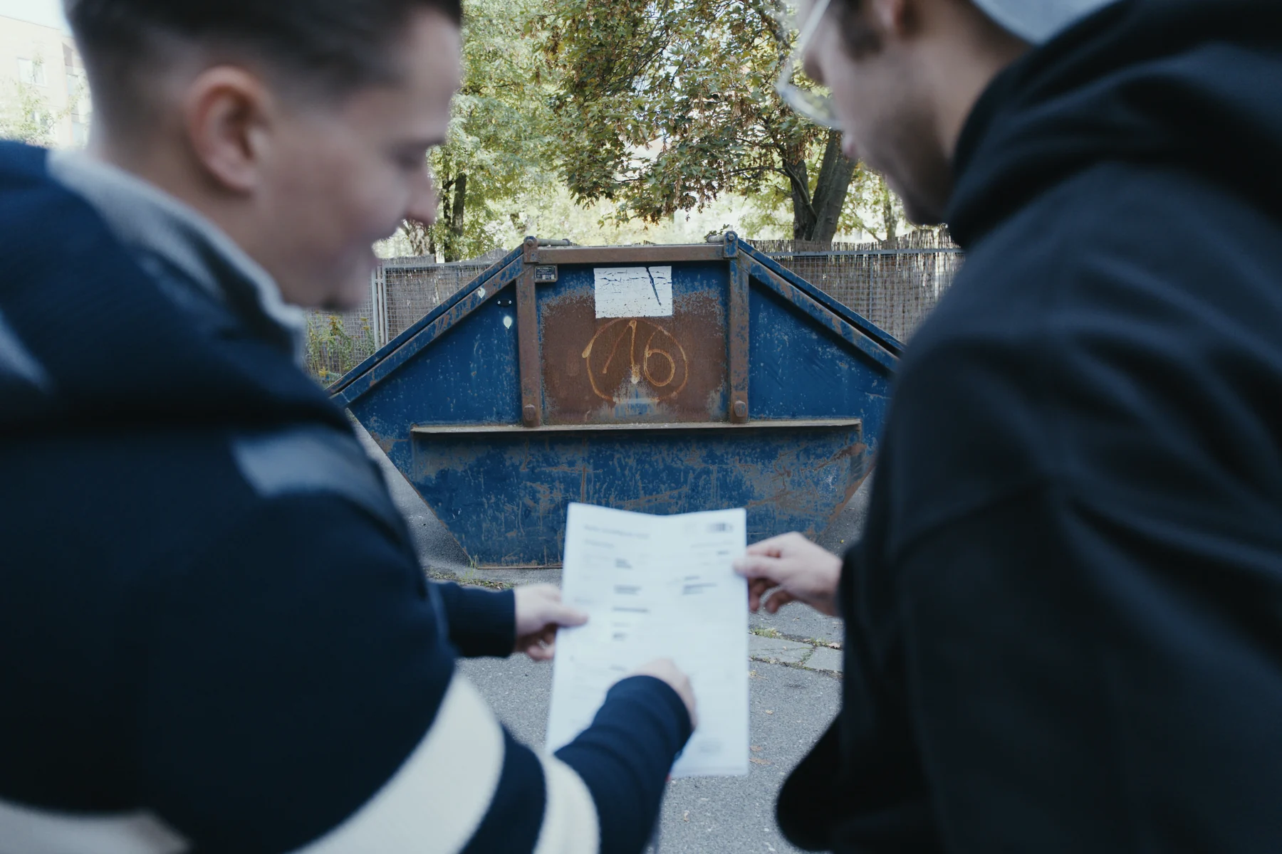 Zwei Mitarbeiter von Sorglos Entrümpeln schauen gemeinsam auf ein Blatt Papier vor einem blauen Absetzcontainer