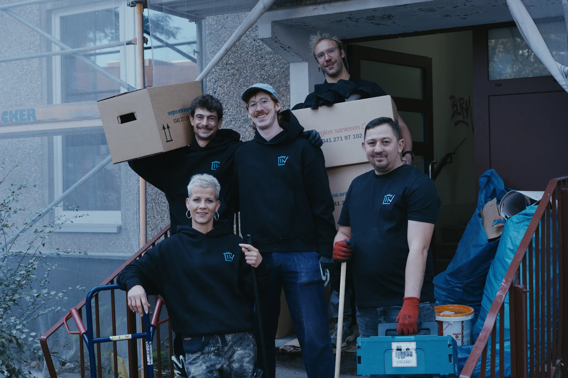 Das Team von Sorglos Entrümpeln vor einem Hauseingang, mit Umzugskartons, Sackkarre, Besen und Werkzeugkoffer