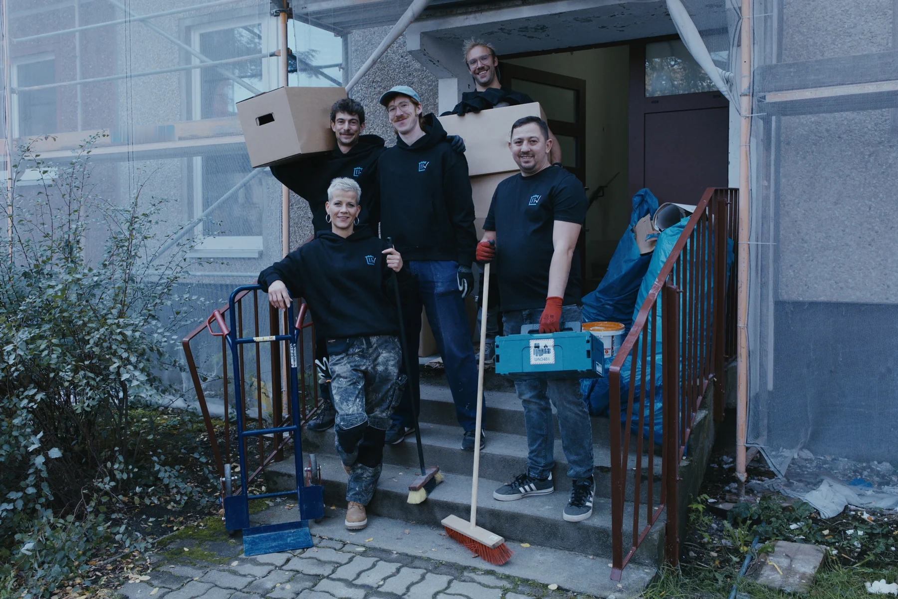 Das Team von Sorglos Entrümpeln auf der Treppe vor einem Plattenbau-Eingang, mit Kartons, Sackkarre, Besen und Werkzeugkoffer