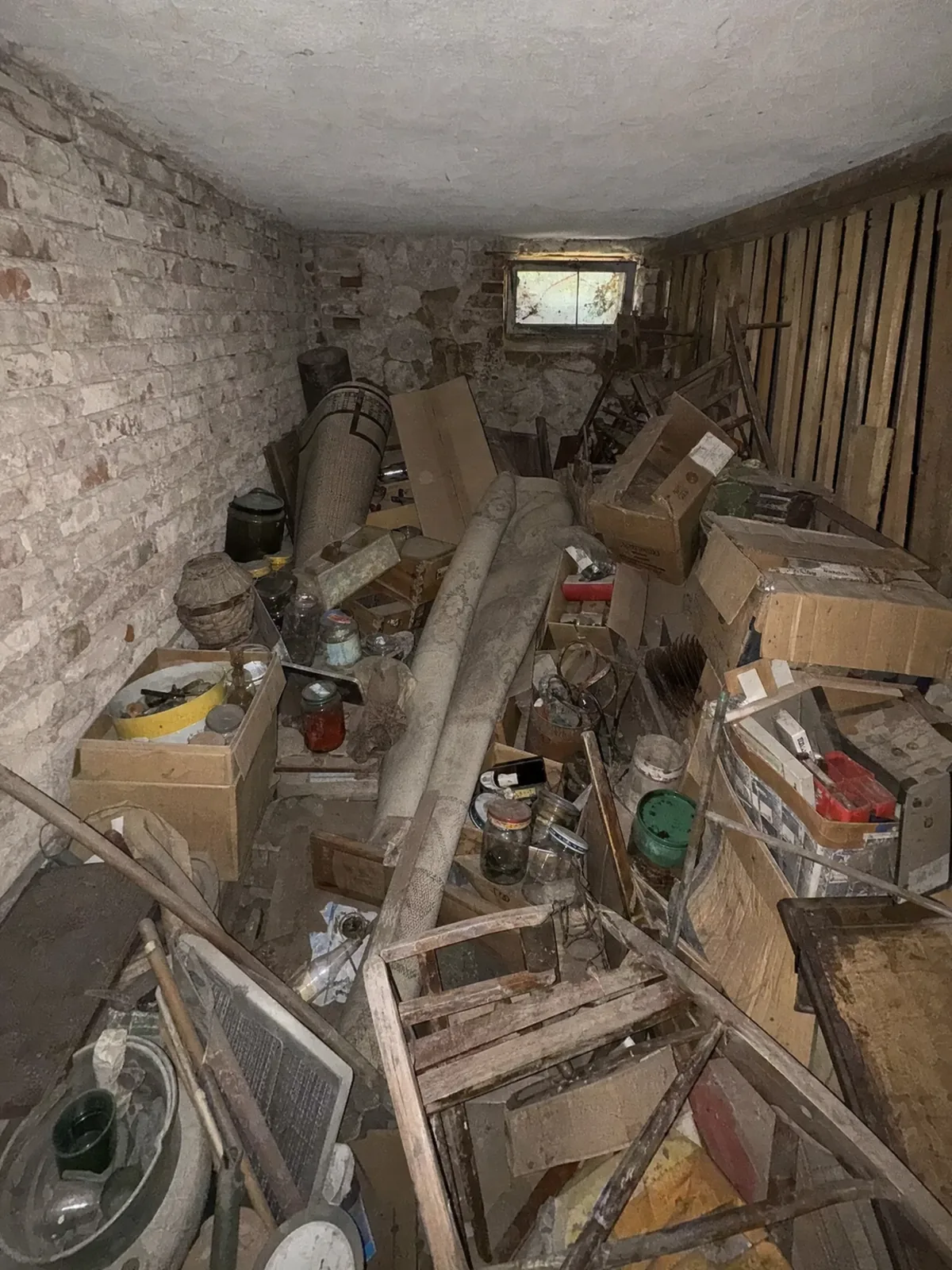 Symbolbild: Kellerraum vor der Räumung, Ziegelwände und Holzlatten, vollgestellt mit Kartons, Gläsern und Hausrat