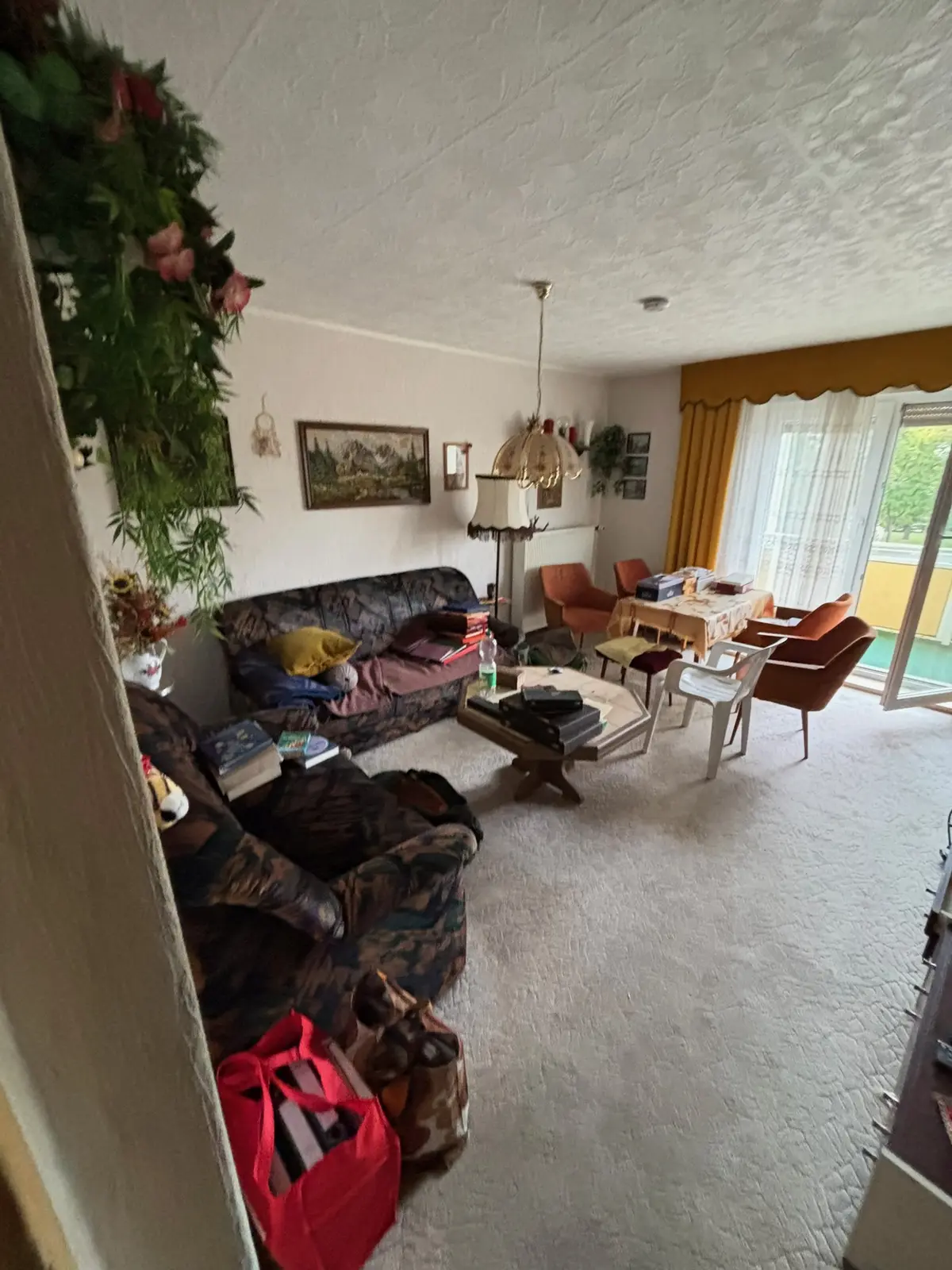 Bewohntes Wohnzimmer vor der Räumung: zwei gemusterte Sofas, Couchtisch mit Büchern, Esstisch und Sessel am Balkonfenster
