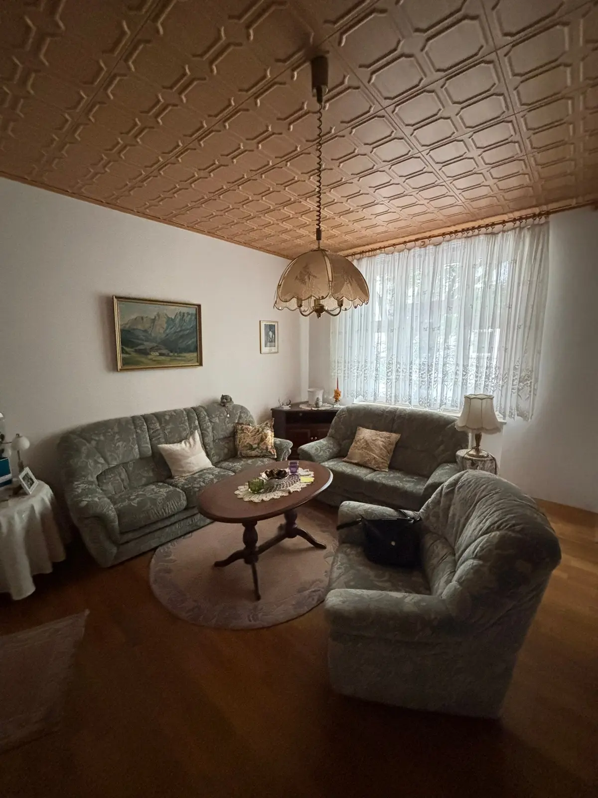 Gepflegtes Wohnzimmer mit Polstergarnitur um einen ovalen Tisch, Gemälde an der Wand, Spitzengardine am Fenster