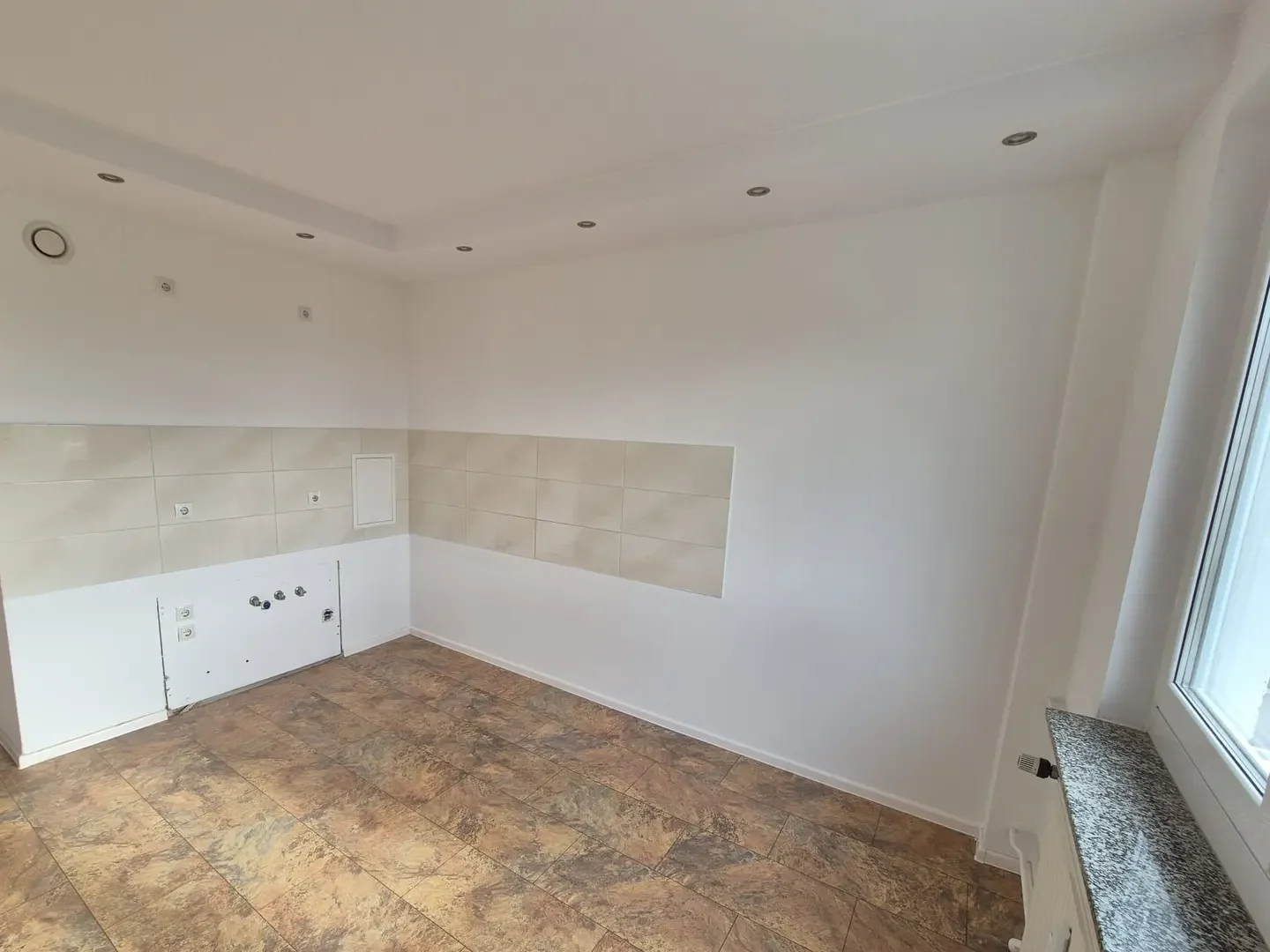 Leergeräumte Küche nach der Räumung: freie Wand mit hellem Fliesenspiegel, offene Anschlüsse für Herd und Spüle, gefliester Boden und Fenster mit steinerner Fensterbank