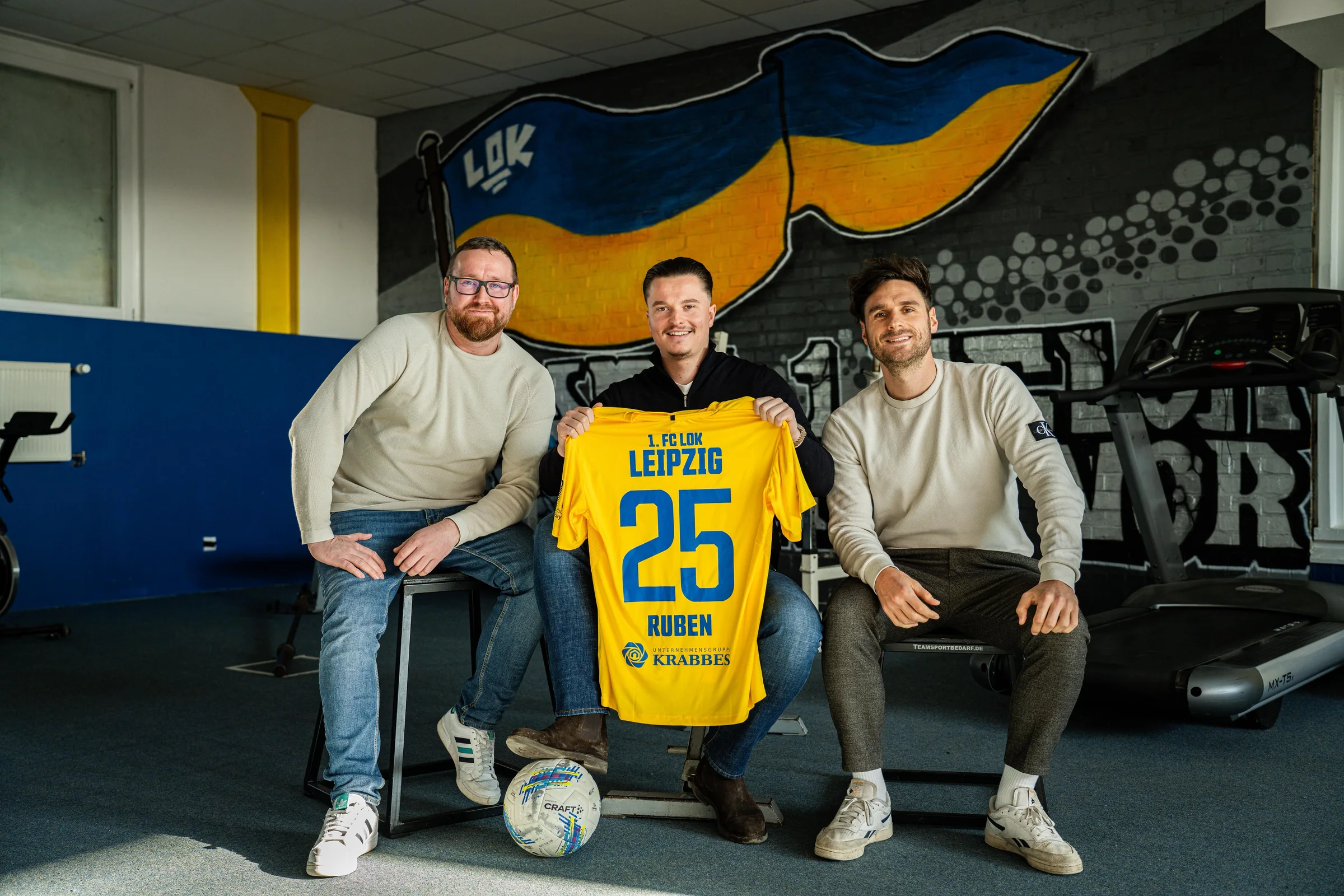 Ruben Bestfleisch hält ein gelbes Trikot des 1. FC Lok Leipzig mit der Nummer 25 und seinem Vornamen, neben ihm zwei Männer, dahinter ein Lok-Wandbild