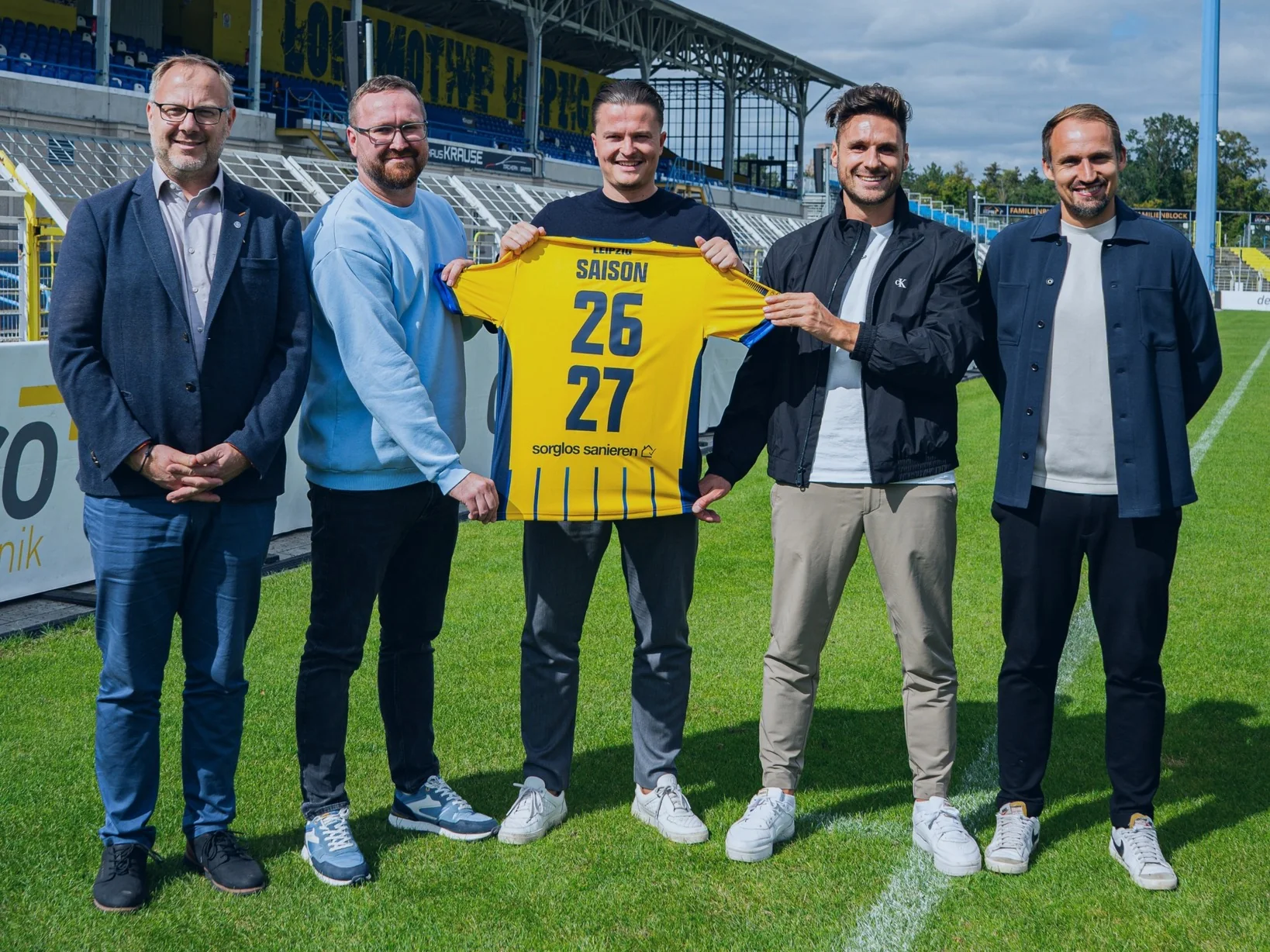 Fünf Männer auf dem Rasen im Bruno-Plache-Stadion, Ruben Bestfleisch in der Mitte hält ein gelbes Lok-Trikot mit der Aufschrift Saison 26/27 und sorglos sanieren